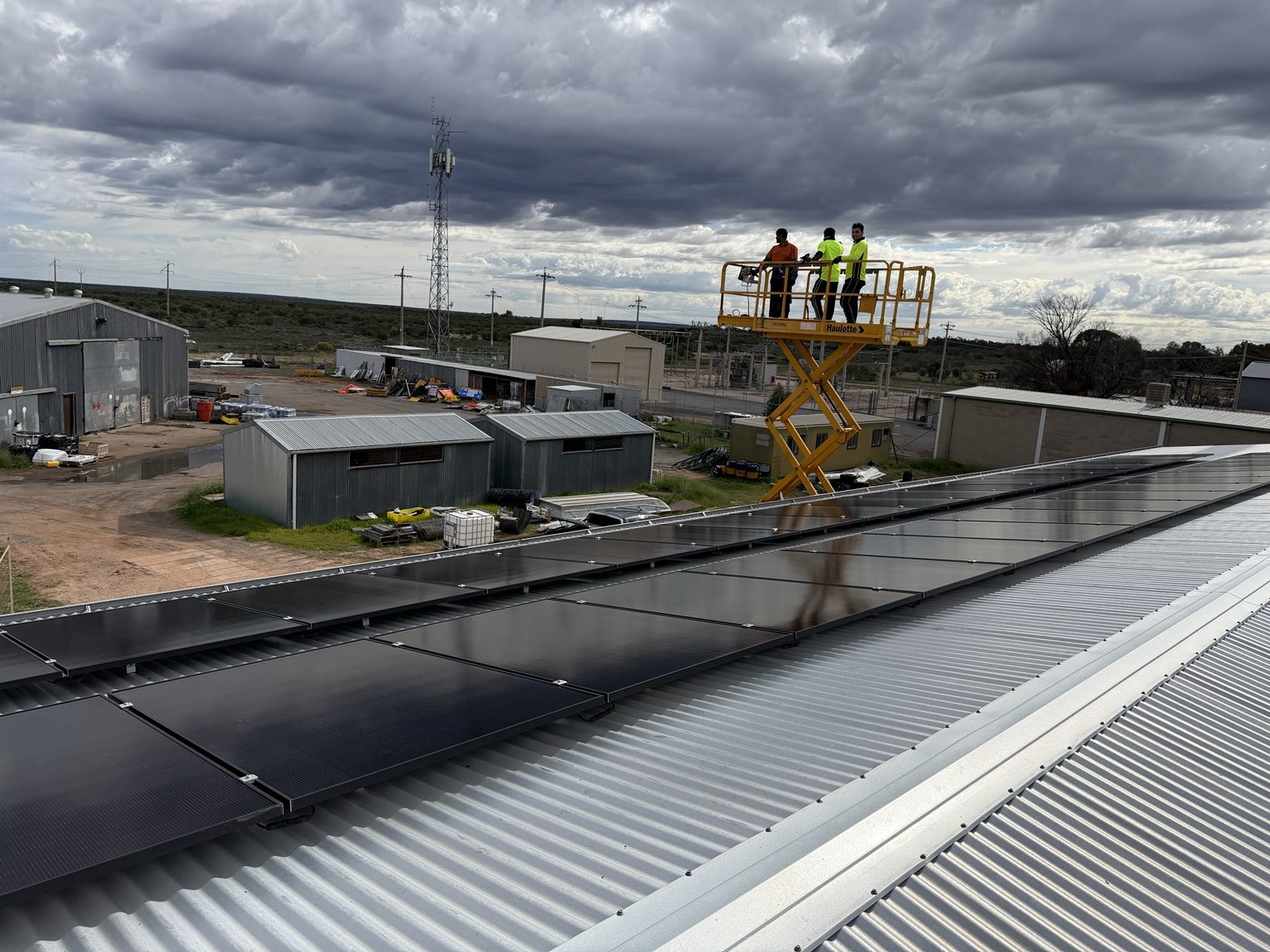 Installers on an elevated work platform beside commercial rooftop solar panels under a cloudy sky