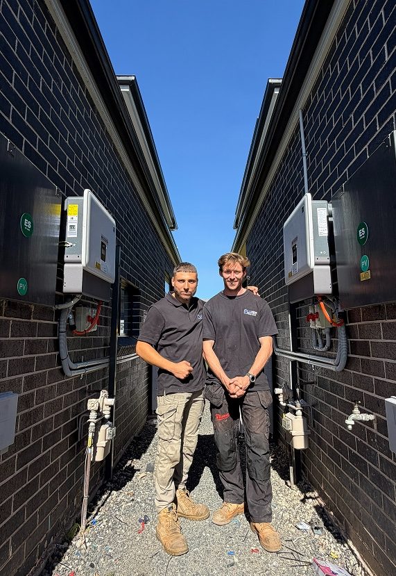 Two Oz Home Energy installers standing between rows of townhouses next to wall-mounted battery units